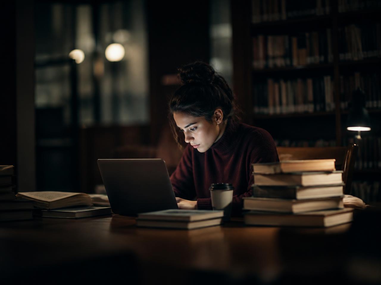 A female student works alone in the evening with a laptop and books in the library